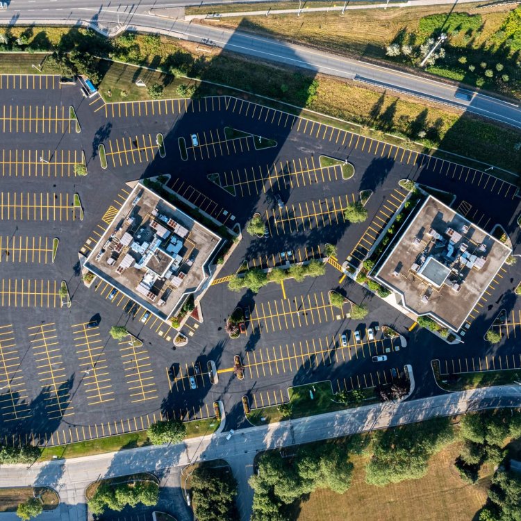 Aerial view of a large, mostly empty parking lot with yellow lines painted on fresh asphalt, surrounding two adjacent buildings. The scene includes greenery, trees, and roads with light traffic. The photo captures the buildings' shadows and neatly arranged paved parking spaces.