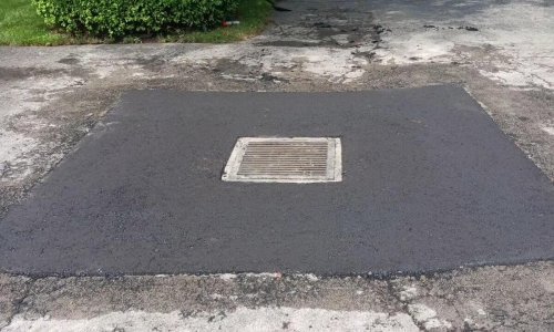 A freshly patched section of road containing a manhole cover sits adjacent to a worn driveway. The surrounding area shows older, tired asphalt. Traffic cones and a few bushes are seen in the background, along with a parked car and residential houses across the street.