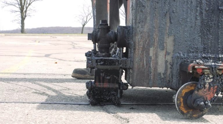 A close-up of road repair machinery applying hot asphalt to cracks in a pavement on a clear day. The machine's wheel and the processed ground around it are visible, showing a gritty texture from the paving mixture being applied to the cracks.