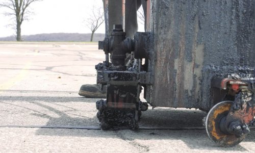 A close-up view of a machine applying crack sealant on an asphalt surface. The machinery's textured, dark sealant is visible, along with its wheels and a part of the operator's boot. The background shows an empty outdoor parking lot with a few bare trees.