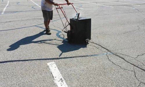 A person in a white shirt and gray shorts pushes a black machine on a cracked asphalt parking lot. There are faint outlines of parking lines on the ground, and the person’s shadow is visible. The image suggests they may be repairing or painting the lines.