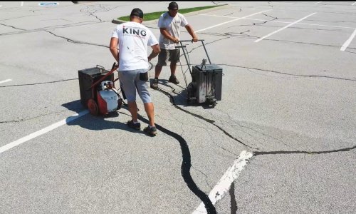 Two workers in shorts and t-shirts, labeled "KING," are sealing cracks in an asphalt parking lot. One operates a machine that applies the sealant, while the other follows with a blower to smooth it out. The otherwise empty lot has visible cracks.