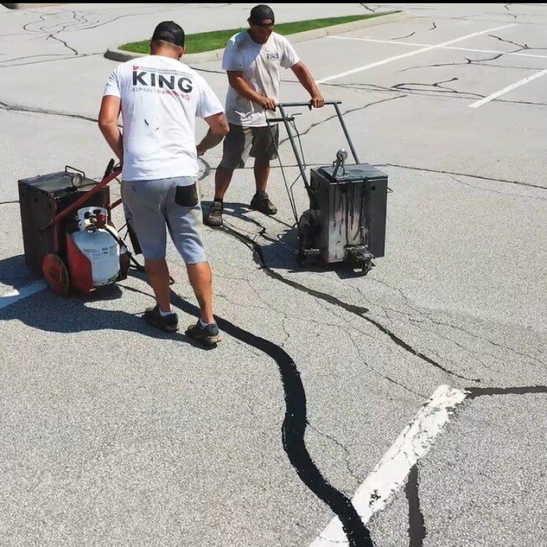 Two workers in shorts and t-shirts, labeled "KING," are sealing cracks in an asphalt parking lot. One operates a machine that applies the sealant, while the other follows with a blower to smooth it out. The otherwise empty lot has visible cracks.