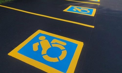 A parking lot with freshly paved dark asphalt has multiple designated handicapped parking spaces. Each spot is marked with a blue square and a yellow outline, featuring a modified accessibility symbol with a dynamic figure in a wheelchair.