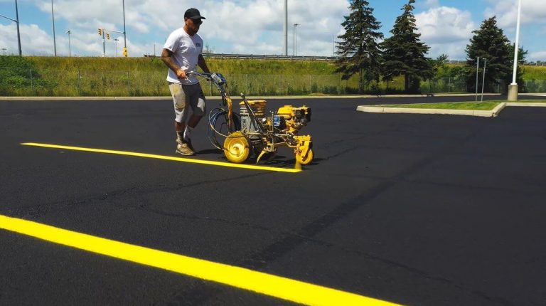 A person operates a line striping machine to paint a bright yellow line on the asphalt of an empty parking lot on a sunny day. The scene features green grass, a few trees, and a blue sky with clouds in the background, showcasing a meticulous paving effort against nature's serene backdrop.