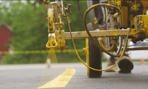 Close-up of a road line striping machine painting a yellow line on asphalt in a parking lot. The machine's nozzle is actively spraying the paint, creating a precise stripe. Background includes blurred trees and a red structure, with yellow caution tape partially visible.