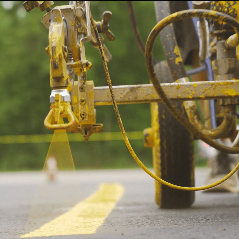 Close-up of a road line striping machine painting a yellow line on asphalt in a parking lot. The machine's nozzle is actively spraying the paint, creating a precise stripe. Background includes blurred trees and a red structure, with yellow caution tape partially visible.