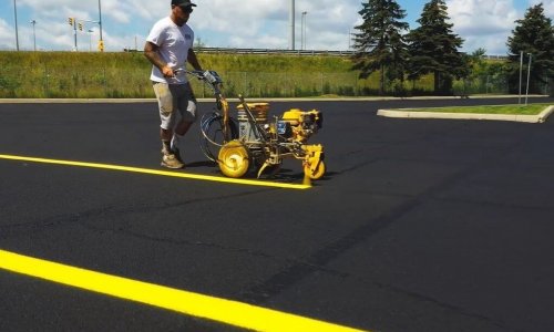 A worker in a white t-shirt and cap operates a yellow line striping machine, painting bright yellow lines on a freshly paved asphalt parking lot. The scene is outdoors on a sunny day with green grass, trees, and a road in the background.