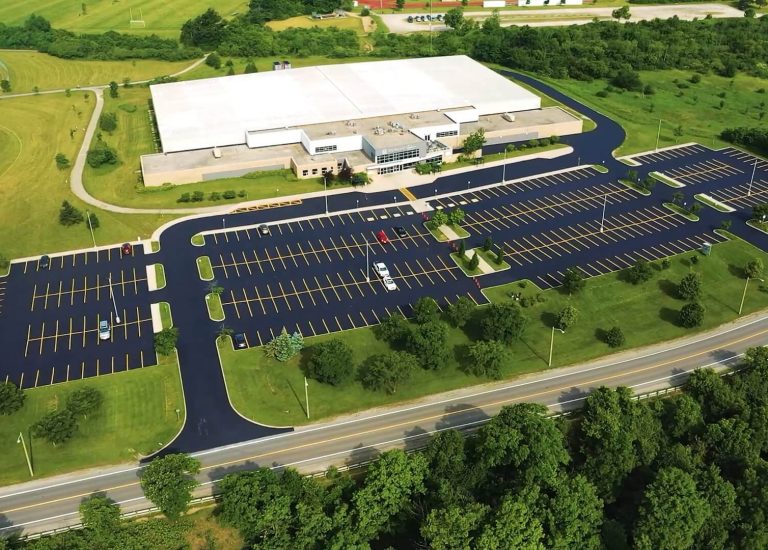 Aerial view of a large, rectangular, white-roofed building surrounded by lush green fields and trees. A newly paved, empty parking lot with marked spaces and smooth asphalt roads lies in front of the building. A two-lane road runs parallel to the parking lot in the foreground.