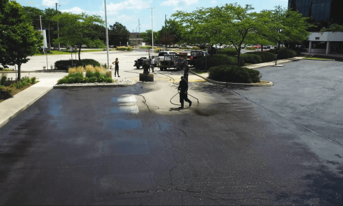 A worker in protective gear is spray sealing an asphalt parking lot. Nearby, a few onlookers and a truck with paving equipment are visible. Trees, neatly landscaped areas, and an adjacent building complete the scene under a clear sky.