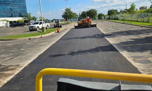 Workers are paving a parking lot with fresh asphalt on a sunny day. A glass office building and several parked vehicles are visible in the background. Trees and a chain-link fence line the right side of the image.