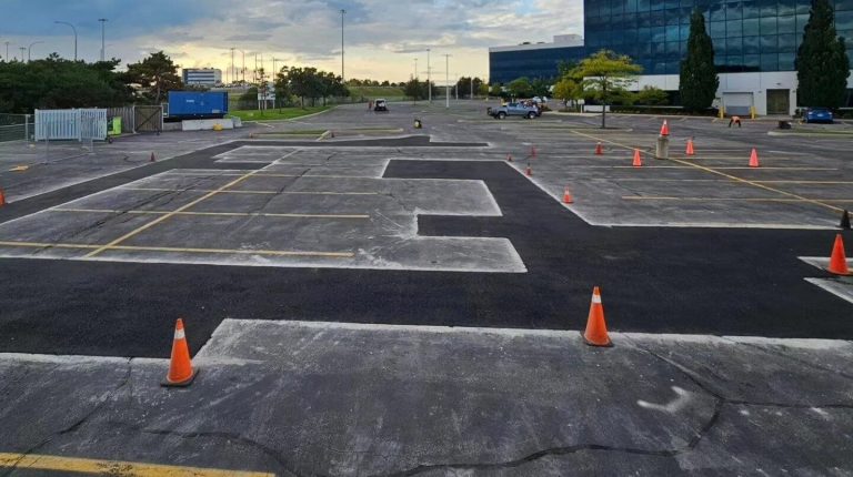 A parking lot undergoing maintenance with traffic cones placed around freshly paved patches of asphalt. The sky is partly cloudy, and a multi-story glass building is visible on the right side. Sparse trees and a security gate are in the background.