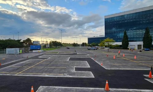 A large, empty parking lot with orange traffic cones arranged in various patterns is visible, its smooth asphalt stretching wide. A modern glass building stands prominently on the right. The sky is mostly cloudy with some patches of blue, and trees and fencing can be seen in the background.