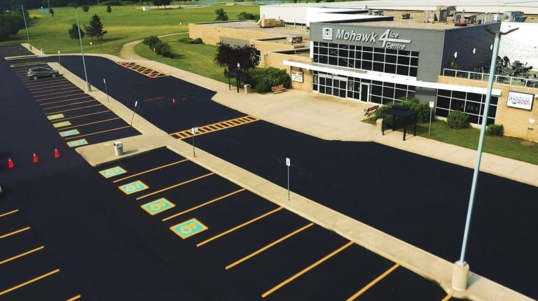 Aerial view of a parking lot with fresh asphalt and several reserved spaces labeled for disabled parking, defined with blue and yellow markings. The lot is adjacent to the modern building labeled "Mohawk 4 Ice Centre," featuring large windows and glass entrance doors.