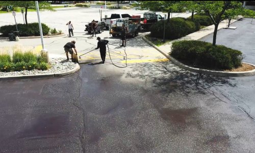 A group of workers using paving equipment to apply a new asphalt surface to a parking lot on a sunny day. Trucks and machinery are parked nearby, and other areas of the lot are still untouched. Shrubs and trees are visible around the perimeter.