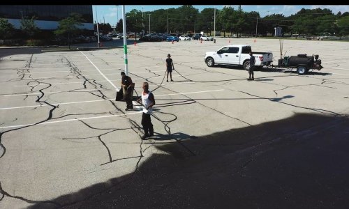 An aerial view shows several workers sealing cracks in a large, empty parking lot. They are applying sealant using tools while a white truck with sealing equipment is parked nearby. Trees and buildings are visible in the background under a clear sky, emphasizing the smooth asphalt surface.