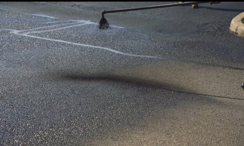 A close-up of a worker applying a sealant to an asphalt pavement with a long-handled tool. The dark, glossy sealant contrasts with the dry, rough texture of the untreated paving. The application process is in progress, with some areas already covered.