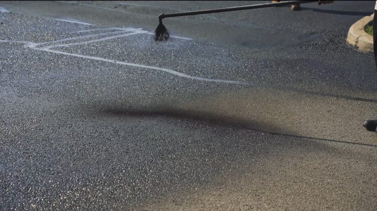 A close-up of a worker applying a sealant to an asphalt pavement with a long-handled tool. The dark, glossy sealant contrasts with the dry, rough texture of the untreated paving. The application process is in progress, with some areas already covered.