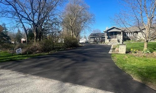 A freshly paved asphalt driveway leads to a single-story house surrounded by leafless trees and grassy areas on a sunny day. The house has a stone mailbox near the entrance and well-maintained landscaping along the driveway.